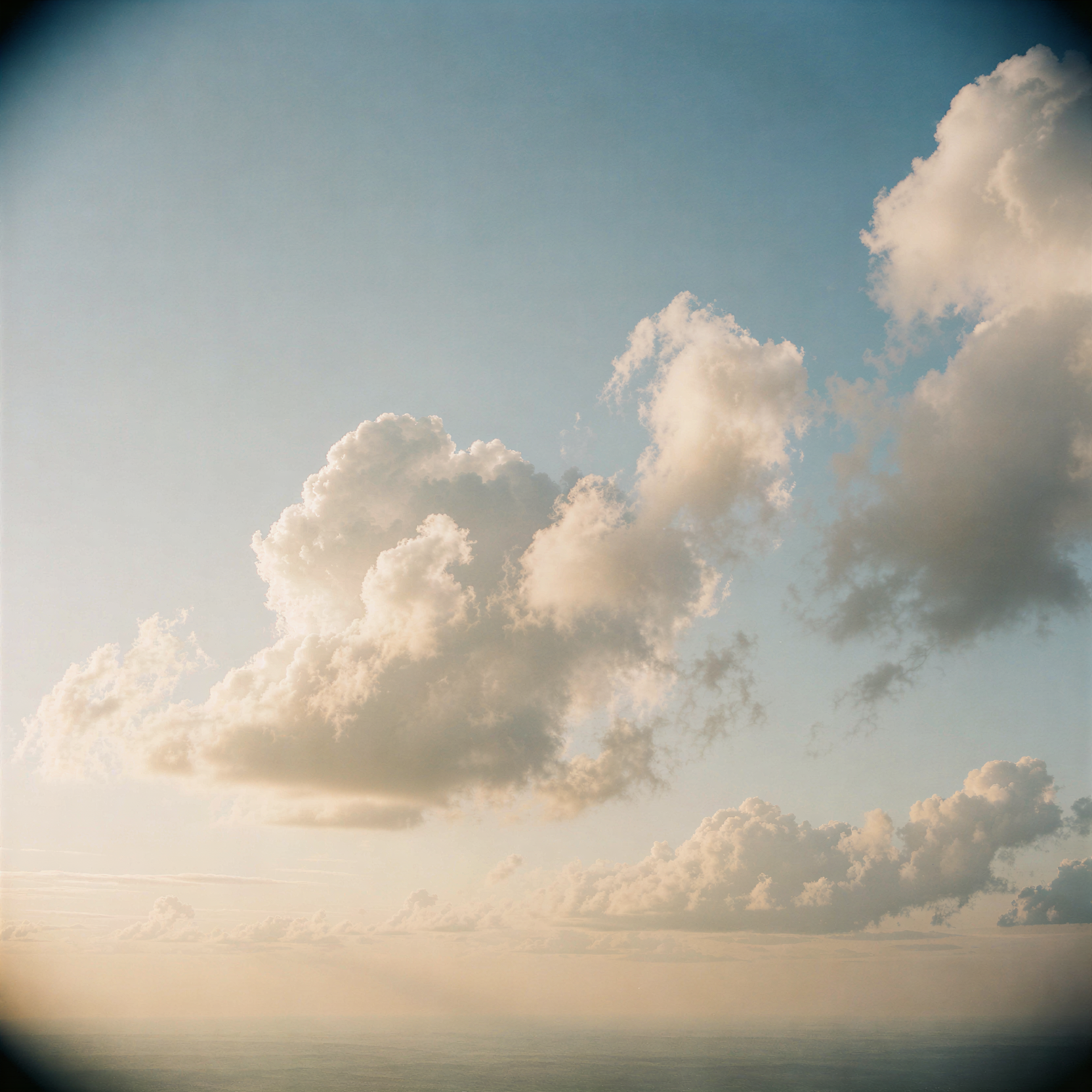a realistic editorial photograph of soft, billowing cumulus clouds drifting slowly across a vast, pale blue sky, illuminated by the warm, golden light of early morning sun. The clouds appear to dance gently in the high-altitude breeze, their edges glowing with a delicate rim light while their undersides hold subtle cool shadows that define their three-dimensional volume. The composition is wide and open, emphasizing the serene scale of the atmosphere above a distant, hazy horizon line. Shot on 35mm Kodak Portra 400 with fine visible film grain and a slight soft vignetting in the corners, capturing the tranquil mood of dawn.
